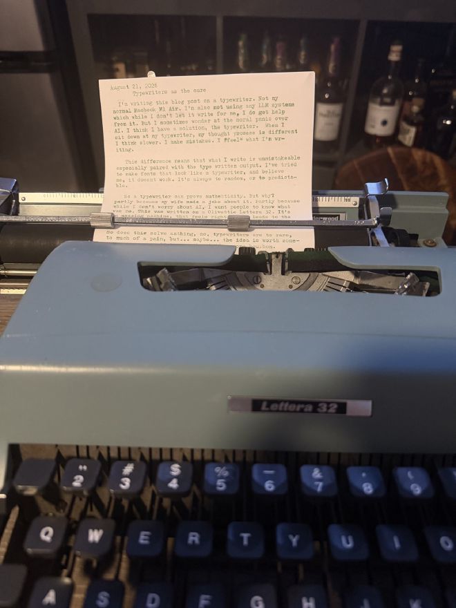 An Olivetti Lettera 32 typewriter in pale blue, the typed manuscript of this post still in the carriage, with shelves of bottles blurred in the background.