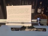 An Olivetti Lettera 32 typewriter in pale blue, the typed manuscript of this post still in the carriage, with shelves of bottles blurred in the background.